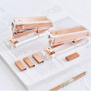 May include: Two rose gold staplers with clear plastic bodies and metal accents. The staplers are on a white surface with several packs of staples.