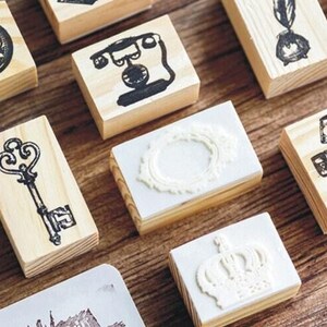 Op de afbeelding: Een verzameling houten stempels met verschillende ontwerpen, waaronder een klok, een telefoon, een sleutel, een kroon en een frame. De stempels zijn allemaal gemaakt van natuurlijk hout en hebben een vintage uitstraling.