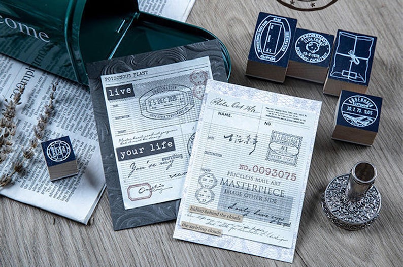 May include: A collection of vintage-style stamps, stationery, and ephemera. The scene includes several stamps with dark blue ink pads, printed paper, and a decorative metal object. The paper has text and stamps, with phrases like "live" and "your life".