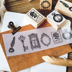 Op de afbeelding: Een set van negen vintage houten stempels met verschillende ontwerpen, waaronder een sleutel, een veer, een raam, een klok, een spiegel en een silhouet van een persoon met een hoed. De stempels zijn gerangschikt op een houten oppervlak met een vel doorzichtig papier waarop de afdrukken van de stempels te zien zijn.