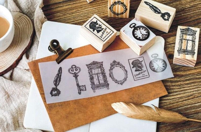 Op de afbeelding: Een set van negen vintage houten stempels met verschillende ontwerpen, waaronder een sleutel, een veer, een raam, een klok, een spiegel en een silhouet van een persoon met een hoed. De stempels zijn gerangschikt op een houten oppervlak met een vel doorzichtig papier waarop de afdrukken van de stempels te zien zijn.