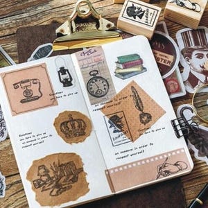 Op de afbeelding: Een open notitieboek met verschillende vintage-stempels en stickers. De stempels tonen afbeeldingen van een kroon, een sleutel, een telefoon, een lamp, een klok, een veer en een pen. De stickers omvatten een vergrootglas, een hand die een pen vasthoudt en een hoed.