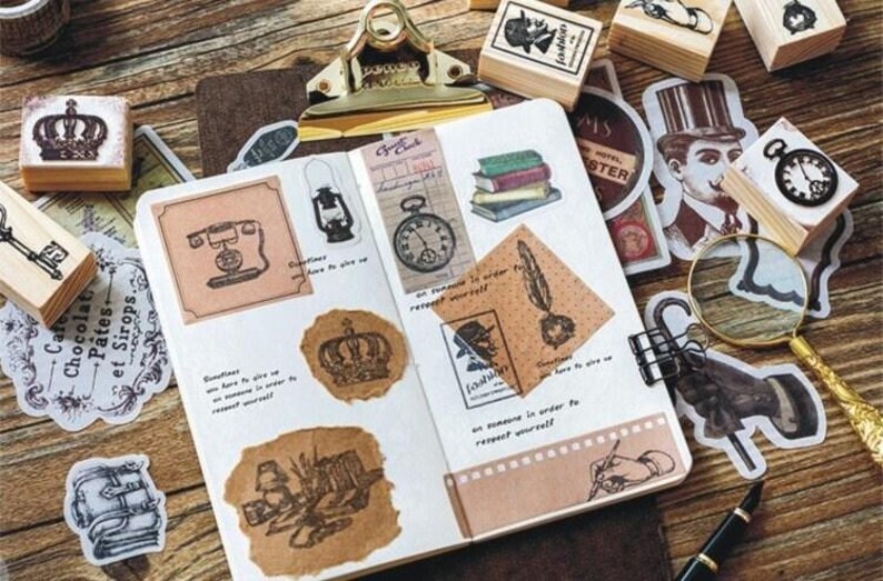 Op de afbeelding: Een open notitieboek met verschillende vintage-stempels en stickers. De stempels tonen afbeeldingen van een kroon, een sleutel, een telefoon, een lamp, een klok, een veer en een pen. De stickers omvatten een vergrootglas, een hand die een pen vasthoudt en een hoed.