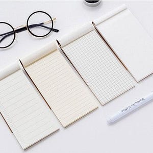 Puede incluir: Cinco pequeños blocs de notas blancos con diferentes patrones de líneas, incluyendo papel en blanco, rayado y cuadriculado. También se incluye un bolígrafo blanco con texto negro.