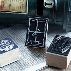 May include: Three wooden stamps with black ink designs sit on a newspaper. One stamp has a package design, another has a circular design with text, and the third has a rectangular design. A dark green mailbox is in the background.
