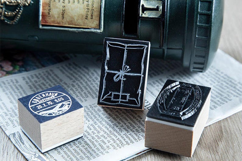 May include: Three wooden stamps with black ink designs sit on a newspaper. One stamp has a package design, another has a circular design with text, and the third has a rectangular design. A dark green mailbox is in the background.