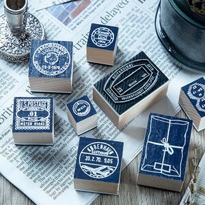 May include: A collection of vintage-style wooden stamps with blue ink imprints, featuring postal and travel-themed designs. The stamps are arranged on a newspaper background, with a silver candlestick and a military-style hat in the background.