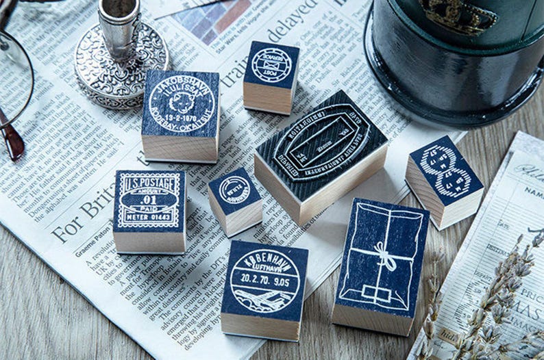 May include: A collection of vintage-style wooden stamps with blue ink imprints, featuring postal and travel-themed designs. The stamps are arranged on a newspaper background, with a silver candlestick and a military-style hat in the background.