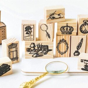 Op de afbeelding: Een set van 12 houten stempels met verschillende vintage ontwerpen, waaronder een vergrootglas, een kroon, een sleutel, een veer, een klok, een raam, een boek, een lamp, een hand die een pen vasthoudt en een silhouet van een persoon. De stempels zijn gerangschikt op een houten oppervlak met een vergrootglas op de voorgrond.