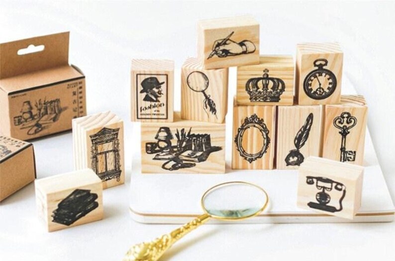 Op de afbeelding: Een set van 12 houten stempels met verschillende vintage ontwerpen, waaronder een vergrootglas, een kroon, een sleutel, een veer, een klok, een raam, een boek, een lamp, een hand die een pen vasthoudt en een silhouet van een persoon. De stempels zijn gerangschikt op een houten oppervlak met een vergrootglas op de voorgrond.