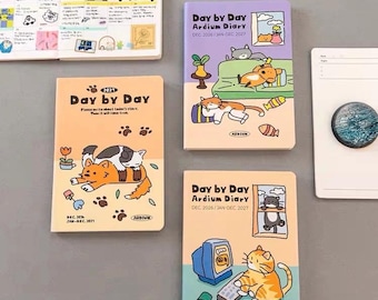 May include: Three rectangular planners with illustrated cat covers. One peach-colored diary features two cats, while the purple and light blue diaries show cats in indoor settings. Each cover displays the text Day by Day Ardium Diary and dates.