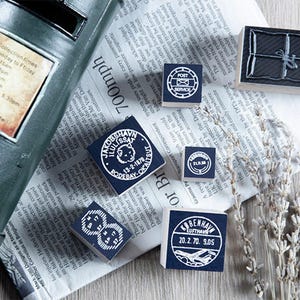 May include: A collection of vintage-style rubber stamps with various designs, including a package, post service emblem, and circular date stamps. The stamps are arranged on a newspaper background, with a mailbox and dried lavender sprigs.