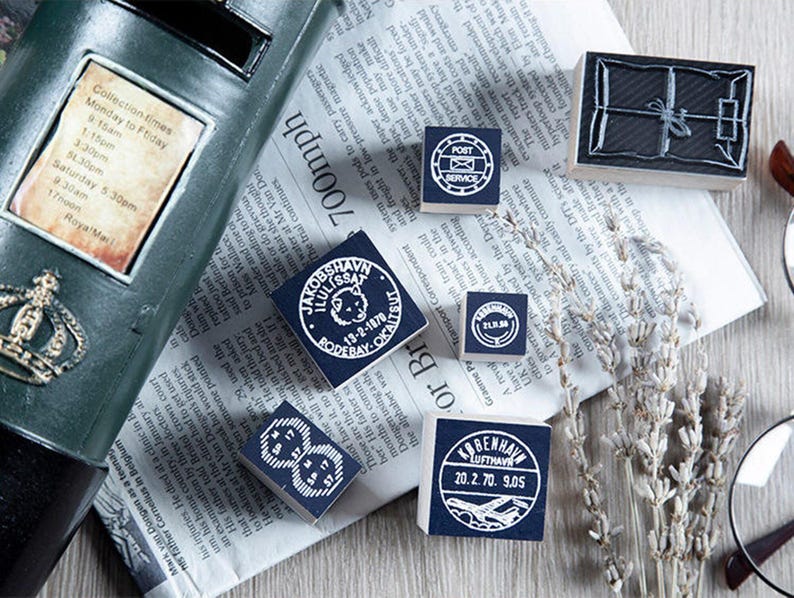 May include: A collection of vintage-style rubber stamps with various designs, including a package, post service emblem, and circular date stamps. The stamps are arranged on a newspaper background, with a mailbox and dried lavender sprigs.