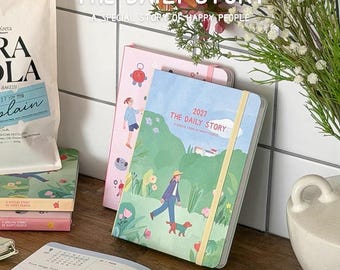 May include: A 2027 planner titled THE DAILY STORY A SPECIAL STORY OF HAPPY PEOPLE featuring a green cover with an illustration of a person walking a dog in a park, secured with a yellow elastic band, alongside an open planner and a pink planner.