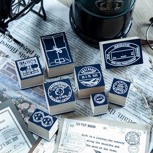 May include: A collection of vintage-style wooden stamps with dark blue ink imprints. Designs include postage, travel, and package motifs. The stamps are arranged on a newspaper background, evoking a nostalgic aesthetic.