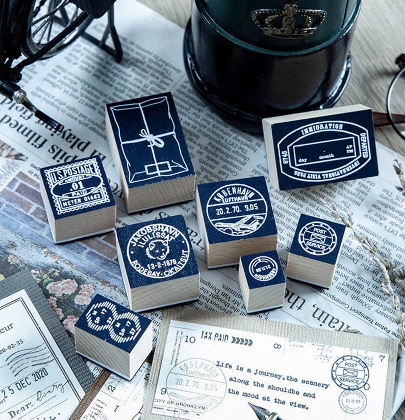 May include: A collection of vintage-style wooden stamps with dark blue ink imprints. Designs include postage, travel, and package motifs. The stamps are arranged on a newspaper background, evoking a nostalgic aesthetic.