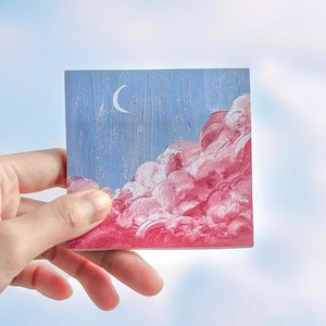 May include: A small, rectangular painting depicting a crescent moon in a blue sky above a pink and white cloud formation.