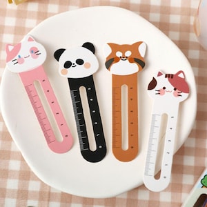 Puede incluir: Cuatro marcapáginas de madera coloridos con diseños de animales. Cada marcapáginas tiene una regla con los números del 0 al 5. Los diseños incluyen un gato rosa, un panda blanco y negro, un panda rojo marrón y blanco y un gato blanco con orejas rojas.