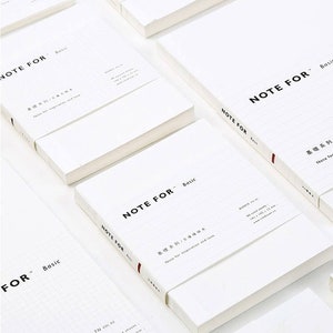 May include: A stack of white notebooks with the text "NOTE FOR - Basic" printed on the cover. The notebooks have lined pages and are designed for note-taking and inspiration.