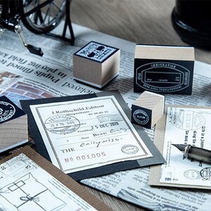 May include: A collection of vintage-style stamps and stationery items. The image features wooden stamps with black ink designs, a fountain pen, and printed documents. The stamps have various designs, including circular and rectangular shapes. The items are arranged on a newspaper background.