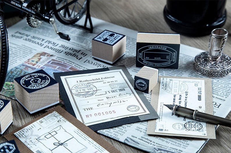 May include: A collection of vintage-style stamps and stationery items. The image features wooden stamps with black ink designs, a fountain pen, and printed documents. The stamps have various designs, including circular and rectangular shapes. The items are arranged on a newspaper background.