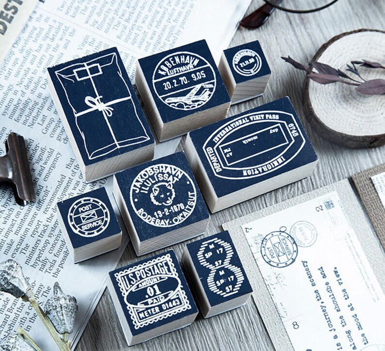 May include: A collection of dark blue and white wooden stamps with various designs, including postal service symbols, travel dates, and a package illustration. The stamps are arranged on a newspaper background.