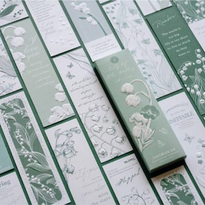 Puede incluir: Un conjunto de marcadores verdes y blancos con diseños de lirio de los valles en relieve y citas inspiradoras. Las citas incluyen "Primavera", "Te amo", "El mundo es demasiado grande", "Eternidad", "Notablemente inefable" y "Solo el valle de los lirios".