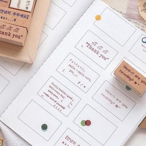 Puede incluir: Una colección de sellos de madera con varias frases y diseños, incluyendo "Gracias", "¿Cómo estás últimamente?" y "fatfatoL smile smile cafe". Los sellos están en una página blanca con algunos otros sellos.