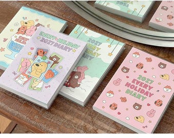 May include: Four rectangular 2027 Every Holiday Diary planners with soft pastel covers. The designs feature cartoon animal illustrations, stars, and clouds. One cover is light purple, one is light green, one is light yellow, and one is pink.