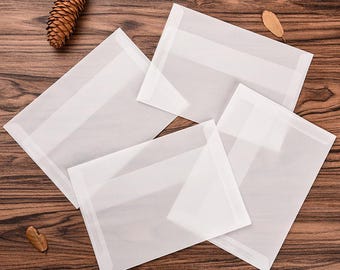 May include: Four white translucent envelopes with a rectangular shape, arranged on a wooden surface. Each envelope has a horizontal band across the middle. A pine cone and a few leaves are also on the wooden surface.