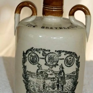 Op de afbeelding: Een vintage keramische kruik met een kurk en twee handvatten. De kruik is gebroken wit met bruine accenten en heeft een zwart-wit illustratie en de tekst "The Greybeard" en "Heather Dew Blended Scotch Whisky".