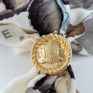 Pode incluir: Um anel de lenço de metal dourado com um design de corda ao redor da borda. O anel apresenta um logotipo em relevo com as letras "CELINE" em um design de duplo "C".