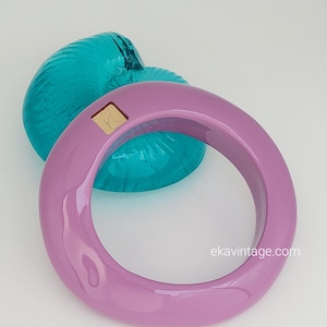 Op de afbeelding: Een gladde, lavendelkleurige armband met een glanzende afwerking en een klein, vierkant goudkleurig metalen accent met de letter K. De armband is geplaatst voor een doorschijnend, talingkleurig, geribbeld object in de vorm van een schelp.