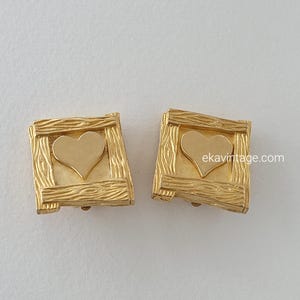 Peut inclure: Une paire de boucles d'oreilles clips dorées avec un motif en forme de cœur à l'intérieur d'un cadre carré. Le cadre a une texture en bois.
