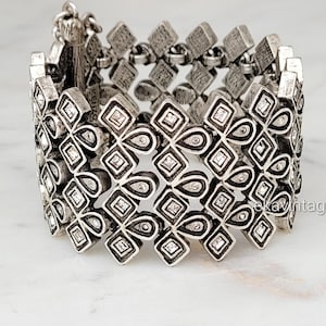 May include: A wide silver-toned metal cuff bracelet featuring a repeating geometric pattern of interconnected diamond and teardrop shapes. Each individual element is accented with a small, clear embedded crystal, creating a textured, link-style design.