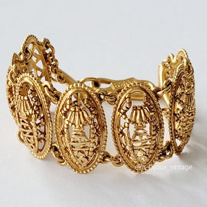 May include: A gold-toned bracelet with a delicate, intricate design. The bracelet is made up of a series of oval-shaped links, each with a detailed pattern. The links are connected by a chain.
