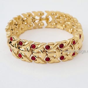 Peut inclure: Un bracelet doré avec un motif tressé, orné de petits accents ronds rouges. Le bracelet est présenté sur une surface blanche, mettant en valeur ses détails complexes et son style vintage.