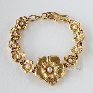 May include: Gold-toned floral bracelet featuring a large flower with a central pearl and smaller flowers with pearls. The bracelet has a clasp closure and is likely made of metal. The design is elegant and suitable for special occasions.