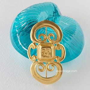 Peut inclure: Une broche dorée au design complexe avec un centre carré orné d'un motif floral. La broche est sur un fond bleu.