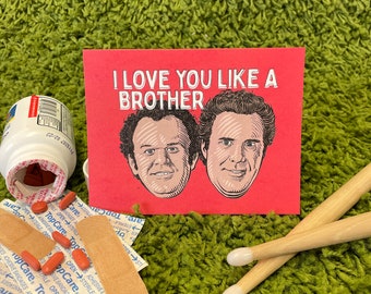 May include: A red card with the text "I LOVE YOU LIKE A BROTHER" in white letters. Two cartoon men with brown hair are pictured on the card. The man on the left has a surprised expression and the man on the right has a serious expression.
