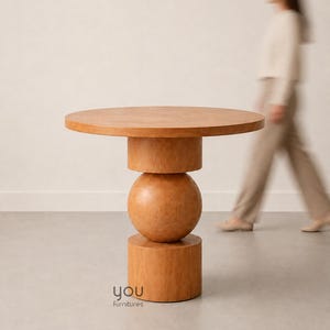 May include: A round wooden table with a light brown finish, featuring a central pedestal base composed of a cylinder, a sphere, and a larger cylinder at the bottom. The text 'you furnitures' is printed in small black letters on the base.
