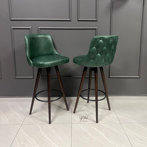 May include: Two green leather bar stools with a tufted design and dark wood legs. The stools have a black metal footrest.