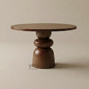 May include: A round wooden table with a smooth, flat circular top and a central pedestal base composed of three stacked, rounded, and bulbous wooden segments. The wood features a medium-brown tone with a visible grain. The text you Furnitures is visible.