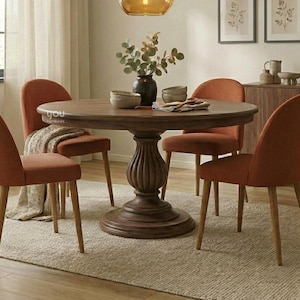 Può includere: Un tavolo da pranzo rotondo in legno con base a piedistallo centrale, circondato da quattro sedie imbottite in tessuto color arancio bruciato con gambe in legno chiaro. Una lampada a sospensione in vetro pende sopra e un vaso con piante è sul tavolo.