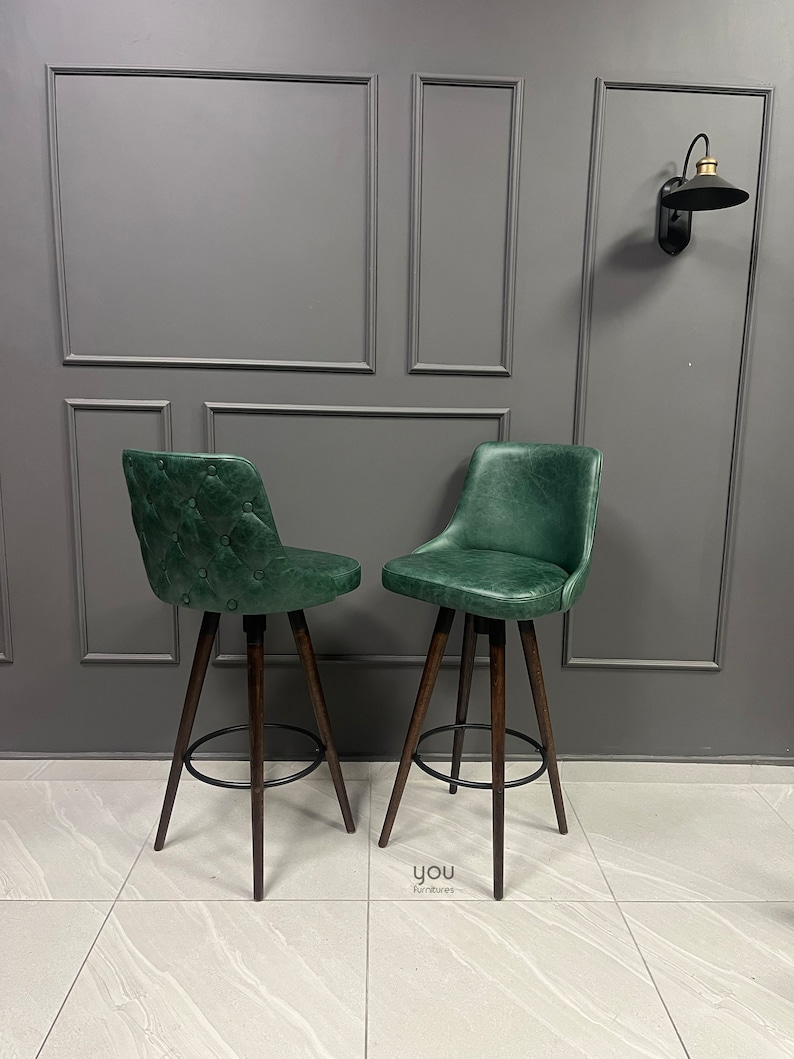 May include: Two green leather bar stools with dark wood legs and a black metal footrest. The stools are set against a grey wall with decorative moulding.