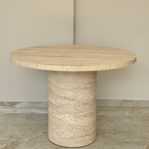 May include: A round travertine dining table with a single pedestal base. The table top and base are a light beige color with natural veining.