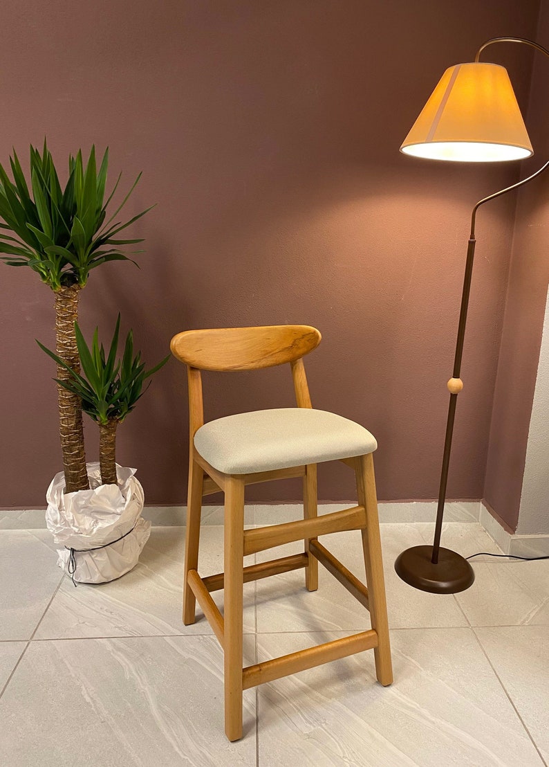 May include: A light wood bar stool with a light gray fabric seat. The stool has a simple design with a curved back and four legs. The stool is in front of a pink wall and a floor lamp with a beige lampshade.