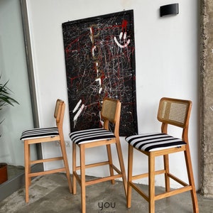 May include: Three wooden bar stools with black and white striped seats and cane backs. The stools are arranged in a row on a concrete floor in front of a black and white abstract painting.