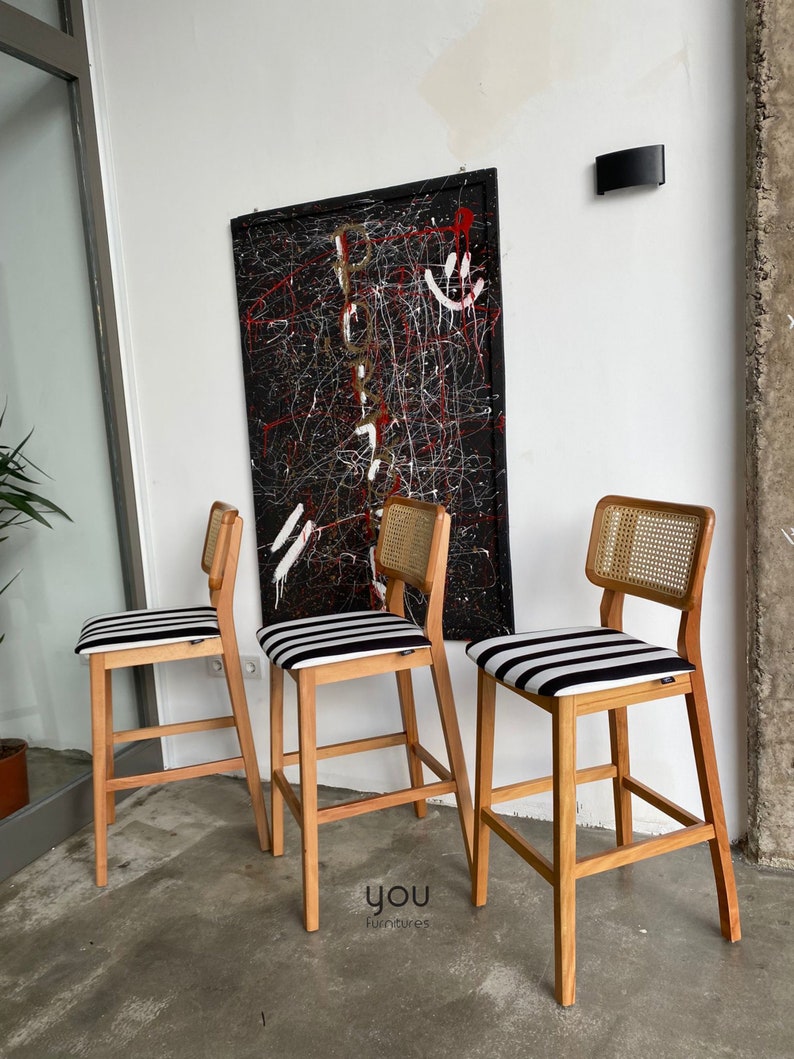 May include: Three wooden bar stools with black and white striped seats and cane backs. The stools are arranged in a row on a concrete floor in front of a black and white abstract painting.