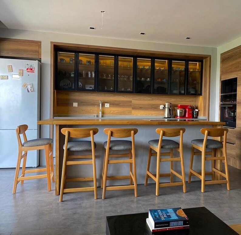 May include: A modern kitchen with a wooden countertop and five wooden bar stools with gray cushions. The countertop is in front of a wall with black framed glass cabinets.